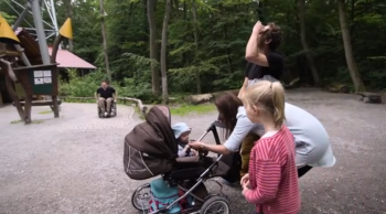 Eine Familie mit Kindern und Kinderwagen auf einem Wanderweg und ein Rollstuhlfahrer im Hintergrund 