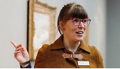 Lächelnde Kunstvermittlerin mit Brille zeigt auf ein Bild im Museum