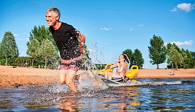 Ein Mann zieht durch das Wasser einen Strandrollstuhl, auf der eine lächelnden Frau sitzt