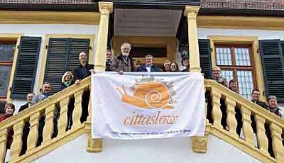 Menschen auf der Freitreppe des historischen Rathauses in Deidesheim mit cittaslow-Fahne. Diese zeigt das Logo mit einer orangenen Schnecke mit kleinen Häusern auf dem Schneckenhaus